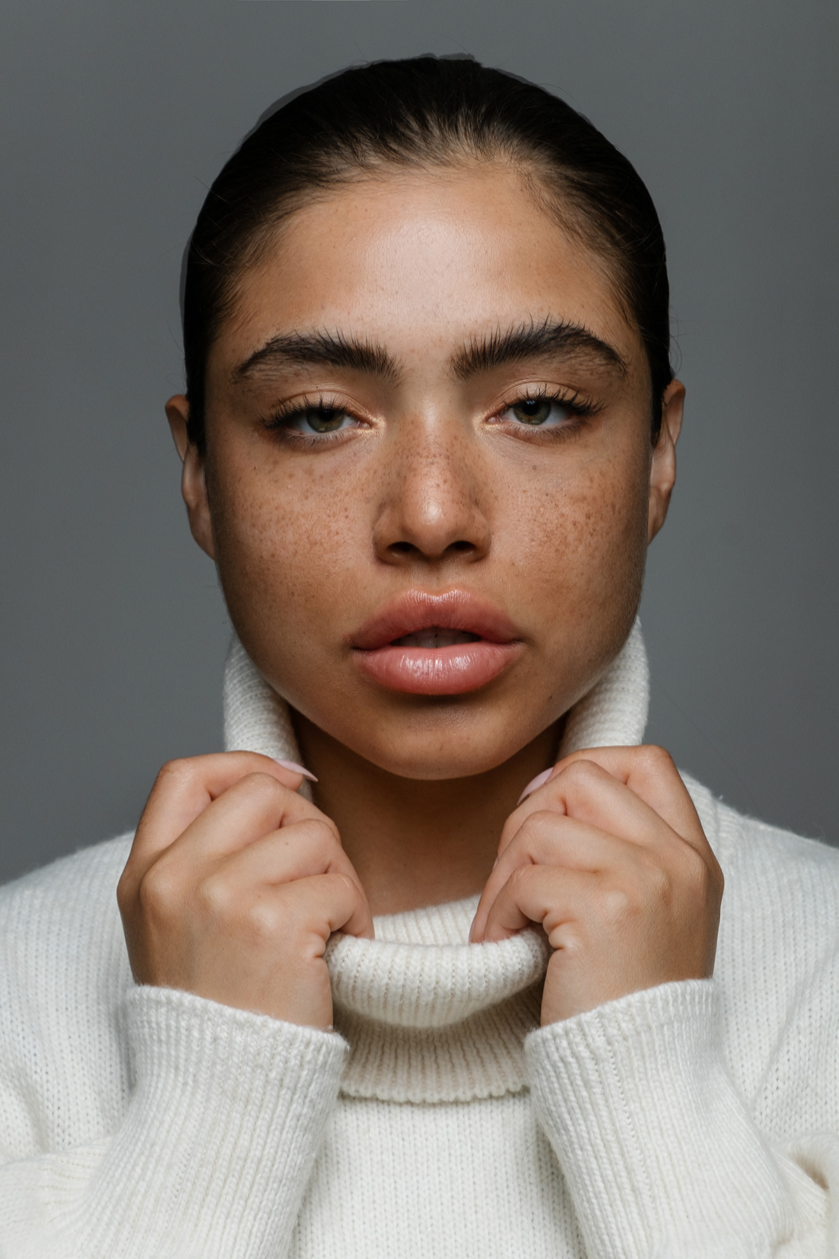 Portrait of a woman with natural makeup, showcasing her freckled complexion, in a cozy white turtleneck sweater, clutching the collar gently, with a neutral background highlighting her serene expression — From a photography studio in Los Angeles