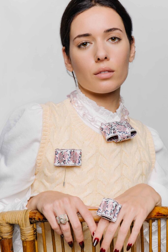 In a portrait capturing the essence of Vidd Studios' Los Angeles eCommerce photography, a woman in a yellow knit sweater overlaid with a white ruffled blouse is adorned with distinctive patterned brooches, her hands elegantly displaying rings that harmonize with her deep red nail polish.