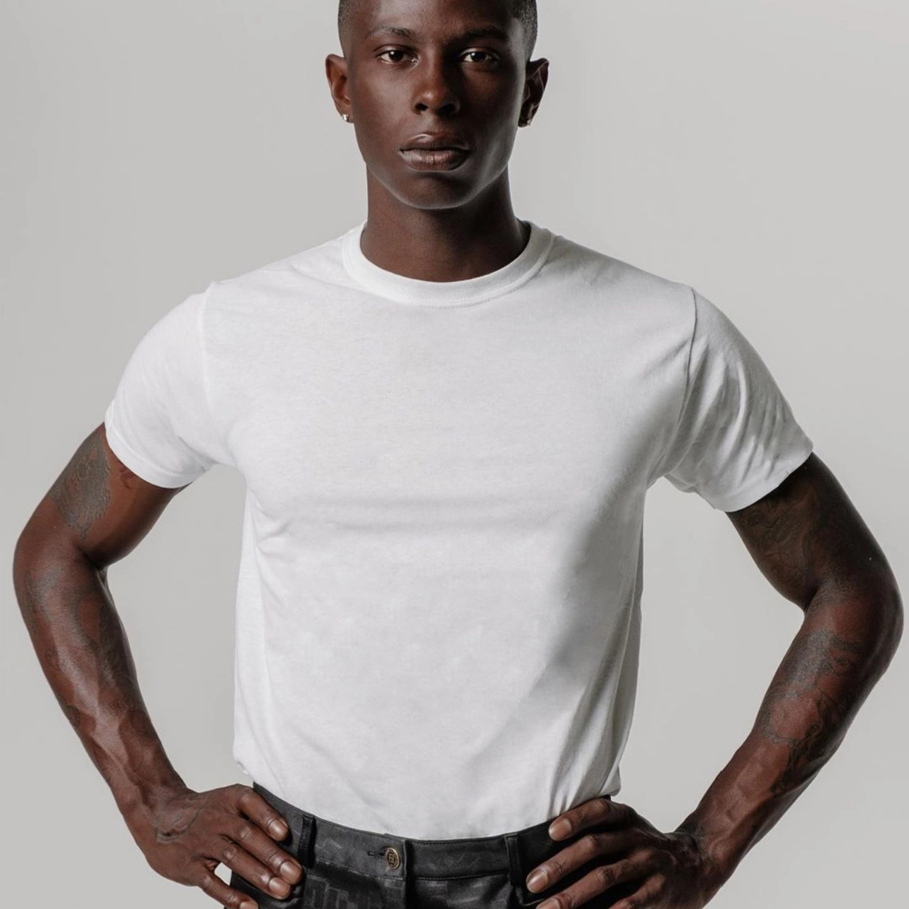 Confident male model with hands on hips wearing a plain white t-shirt and black pants, showcasing a casual yet bold look against a light grey background — A Los Angeles photography studio portfolio